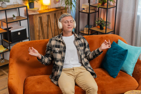 Keep calm down, relax. Man with gray hair listening music, breathes deeply eyes closed meditating with concentrated thoughts peaceful mind at home apartment indoors. Young guy sitting in room on couchの写真素材
