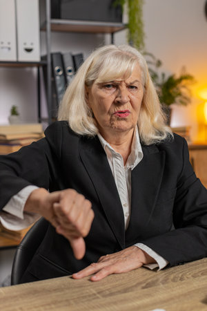 Upset elderly businesswoman sitting at home office showing thumb down sign gesture, expressing discontent, disapproval, dissatisfied bad work. Displeased serious freelancer grandma in suit.の写真素材
