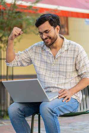 Handsome young man using laptop computer celebrating win good message news, lottery jackpot victory, giveaway online outdoors. Happy Indian guy tourist sitting in city cafe terrace. Town lifestyles.の写真素材