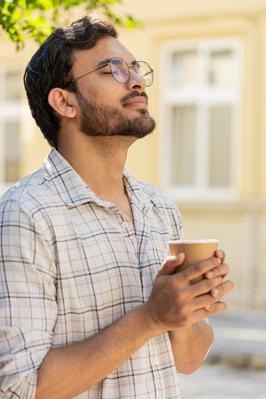 Happy Indian man enjoying morning coffee hot drink and smiling. Relaxing, taking a break. Hispanic young guy standing on urban city center street, drinking coffee to go. Town lifestyles outside.の写真素材