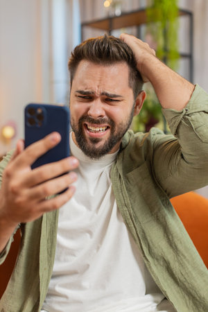 Upset displeased Caucasian man using smartphone sitting on sofa couch in living room. Young adult guy in casual clothes touching head becomes surprised by sudden results or bad news.の写真素材