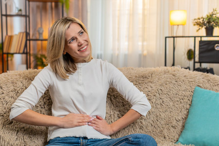 Middle-aged woman holds belly with pain on home sofa, clearly expressing discomfort and tension....