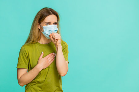 Middle-aged woman coughs and carefully puts on medical mask, showing concern about flu, cold, or infection risk. Girl isolated on blue background expresses safety awareness and illness prevention.の写真素材