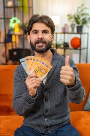 Man at home counts cash banknotes plans travel budget thinks where to spend salary, earnings, savings. Guy holds money stack dreams of trip spend plan feels very happy confidentの写真素材