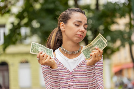 Poor Caucasian young woman holding one dollar banknotes outdoors. Lack of money to buy purchase something in store. Financial crisis. Bankruptcy. Poverty and destitution. Girl on urban city streetの写真素材