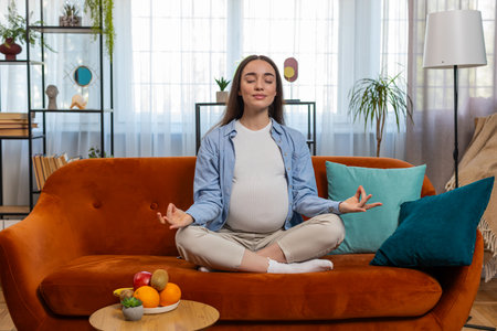 Calm pregnant charming woman sitting in lotus position while meditating on comfy sofa in living room at home. Concept of health care, pregnancy and breathing practice. Happy lady prepares to be motherの写真素材
