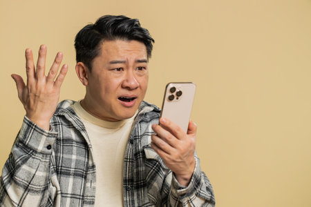 Upset sad Asian man use smartphone typing browsing, play game, loses, surprised by sudden lottery results bad fortune loss fail unlucky news. Chinese middle-aged guy isolated on beige backgroundの写真素材