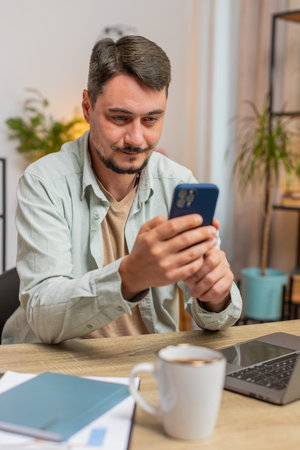 Happy young Caucasian man sitting at table using mobile phone smiling at home apartment. Guy freelancer texting share messages on smartphone social media applications online watching movie. Vertical.の写真素材