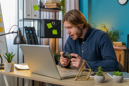 Excited Caucasian businessman celebrating victory in video game on laptop spending leisure time at home office. Happy freelancer guy boy playing video games holding controller. Relaxing taking a breakの写真素材