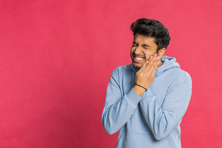 Dental problems. Indian Hindu man touching cheek, closing eyes with expression of terrible suffer from painful toothache, sensitive teeth, cavities. Arabian young guy isolated on red backgroundの写真素材