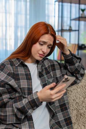 Young woman at home holding smartphone and looking at screen with sad disappointed face after bad news. Caucasian redhead girl on sofa feeling upset, emotional reaction to message or unexpected resultの写真素材
