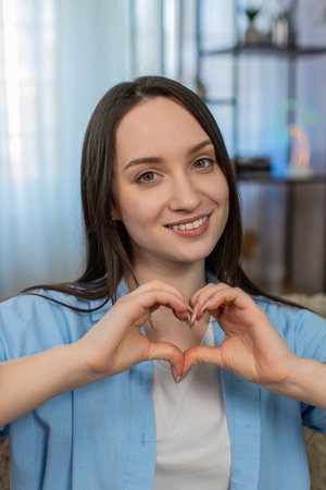 Cheerful young woman at home sitting on sofa showing heart gesture, happily expressing positive...