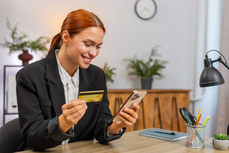 Young businesswoman at home office taps smartphone shows credit card smiles at successful payment. Freelancer girl at table nods happy with transaction enjoys secure online checkout and retail gain.の写真素材