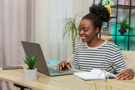 African American Black woman working indoors at home. Young girl sitting on sofa using laptop,...