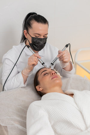 Cosmetologist doctor makes woman micro currents face forehead therapy. Girl skin care anti-age spa...
