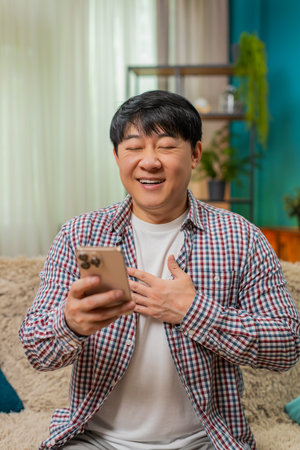 Asian man sitting on home sofa reads good love news on smartphone sighs with relief hand on chest, eyes closed. Chinese guy shows calm thankful expression relaxed smiling gently after positive messageの写真素材