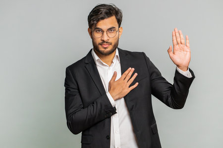 I swear to be honest. Sincere responsible businessman raising hand to take oath, promising to be honest, to tell truth, keeping hand on chest, justice. Man guy on gray studio backgroundの写真素材