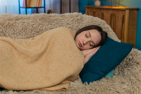Young woman at home falling asleep on sofa wrapped comfortably in blanket, placing head gently on...