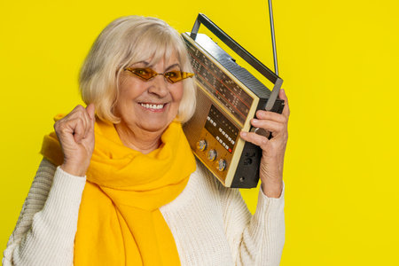 Senior old woman using retro tape record player to listen music, disco dancing favorite track, having fun, entertaining, fan of vintage technologies. Mature grandmother pensioner on yellow backgroundの写真素材