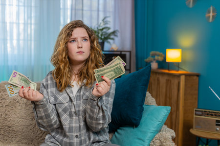 Young woman at home holding cash dollars in hands counting money and thinking about finances with...