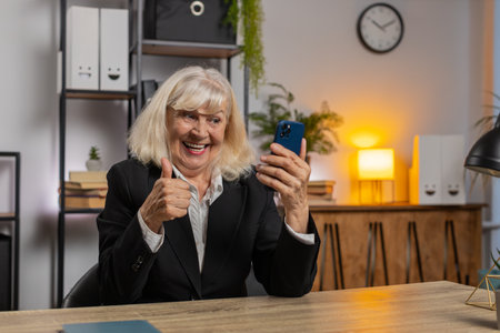 Happy mature woman using smartphone typing browsing yes celebrating success victory winning lottery jackpot goal achievement play game good positive news triumph. Elderly woman at home office table.の写真素材