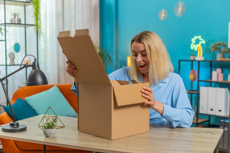 Happy blond businesswoman unpacking delivery parcel sits at desk in apartment. Smiling satisfied...