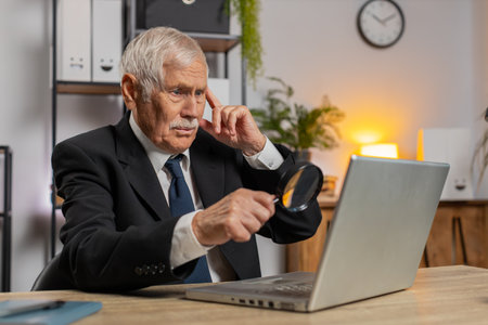 Bored investigator researcher senior businessman working at home office holding magnifying glass looking at laptop screen, searching, analyzing. Caucasian grandpa professional freelancer in suit.の写真素材