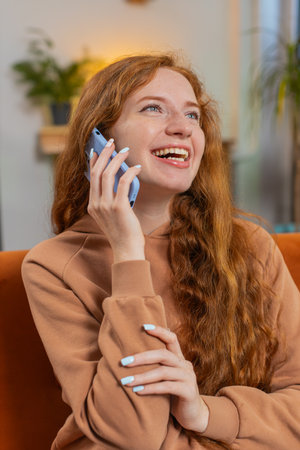 Smiling young Caucasian girl making phone conversation with friends sitting on sofa couch at home in...