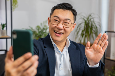 Happy excited Asian middle-aged businessman blogger influencer making phone conversation call with social media followers at office workplace working on laptop. Freelancer Chinese man in formal suitの写真素材