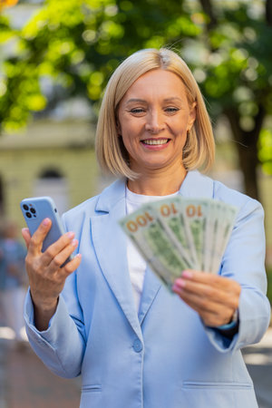 Caucasian mature businesswoman standing on city street showing earnings winning at betting cash money with mobile phone in hands. Middle-aged woman in a suit outdoors with smartphone and pack dollars.の写真素材