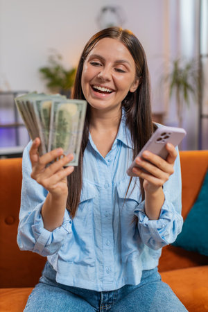 Smiling happy rich Caucasian young woman counting money cash, use smartphone calculate domestic bills sitting on home couch. Girl satisfied of income salary and saves money for planned vacation, giftsの写真素材