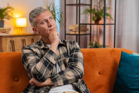 Thoughtful clever Caucasian man with gray hair rubbing chin, looking aside with pensive expression, pondering a solution doubting question at home apartment indoors. Young guy sitting in room on couchの写真素材