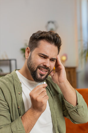 Man suffering from headache taking pill, wellness and treatment in living room, care and relief....
