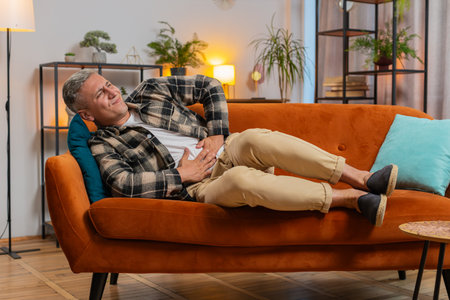 Young Caucasian man lying on sofa feeling sudden strong abdominal stomach ache, gastritis problem. Guy having symptom diarrhea indigestion peptic ulcer pancreatitis at home apartment room.の写真素材