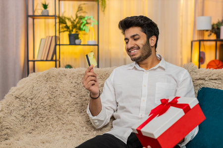 Excited man at home holding credit card, making an online purchase, waiting for payment confirmation. Young guy on sofa, surprised and excited after receiving unexpected gift deliveryの写真素材