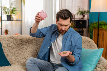 Young man at home shaking a piggy bank, watching coins fall out while checking savings. Caucasian...