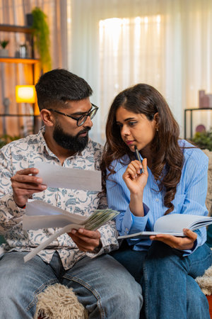 Indian couple man and woman sitting on home sofa, calculating bills and expenses with money and notebook, showing concern. Family managing home budget as girl writes and man reacts with frustration.の写真素材