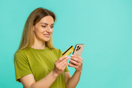 Middle-aged woman clicks order button while holding smartphone and credit card, showing digital payment moment with confidence. Adult girl on blue background, buying goods online with focused emotion.の写真素材