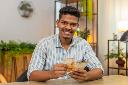 Indian man at home holds cash counts bonus and salary smiles plans invest and grow savings. Guy at table spreads banknotes checks wallet updates budget sets plan for safe money goalsの写真素材