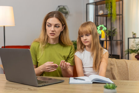 Mother and daughter at home join laptop video call to teacher show notebook give short homework update. Woman and girl at table nod smile listen answer question mark progress note and plan next taskの写真素材