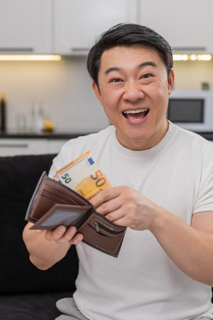 Asian man at home holds cash fan laughs makes victory sign after count and feels proud. Guy on sofa...