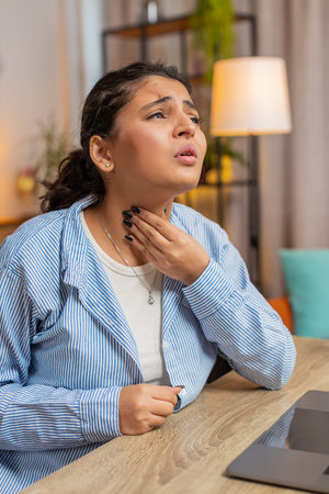 Indian woman freelancer feeling unwell sick ill suffering from sore throat hurt swallowing pain touches neck coughing working at home office workplace. Female girl has a cold, flu, virus. Vertical.の写真素材