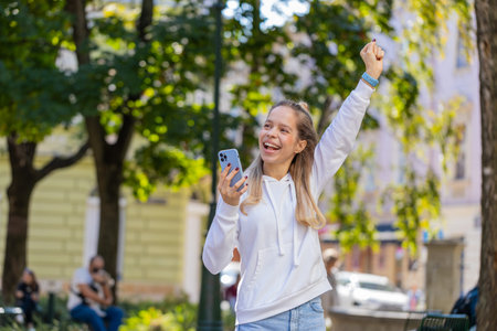 Happy Caucasian young woman using smartphone celebrating win good message news, lottery casino app jackpot victory, online outdoors. Attractive female tourist teenager clenching fists on city street.の写真素材