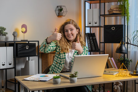 Like. Smiling Caucasian businesswoman looking approvingly at camera showing double thumbs up sign positive something good great news positive feedback. Happy blonde girl using laptop at home officeの写真素材