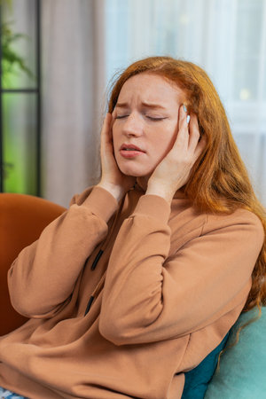 Sick redhead woman rubbing temples to cure headache problem, suffering from tension and migraine stress grimacing in pain at home. Young girl indoors in room on couch. Verticalの写真素材