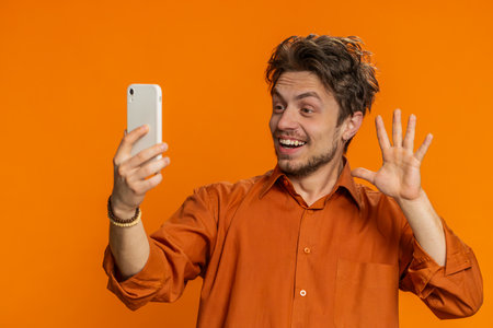 Happy Caucasian man blogger taking portrait selfie on smartphone, streaming vlog, communicating record video call online with social media subscribers. Smiling young guy isolated on orange backgroundの写真素材