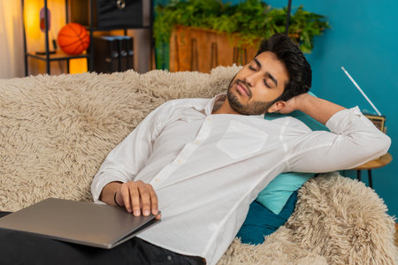 Young man at home, exhausted from work, falling asleep on the sofa with a laptop on his lap. Guy tired after long hours of work, resting on the couch, overwhelmed and drainedの写真素材