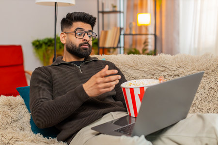 Man at home watches laptop while eating popcorn, laughing during movie scene with joyful face. Guy...