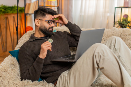 Indian man at home reacts to Bitcoin BTC drop, closing laptop and holding coin with unhappy look. Arabian guy on sofa expresses disappointment and stress, emotionally affected by falling crypto valueの写真素材