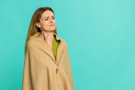 Middle-aged woman wrapped in blanket, touches throat opens mouth showing pain while swallowing, sign of cold or flu. Young adult girl isolated on blue background expresses discomfort illness symptomsの写真素材